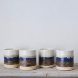 Four ceramic cups with blue and brown stripes on a wooden surface against a light gray background