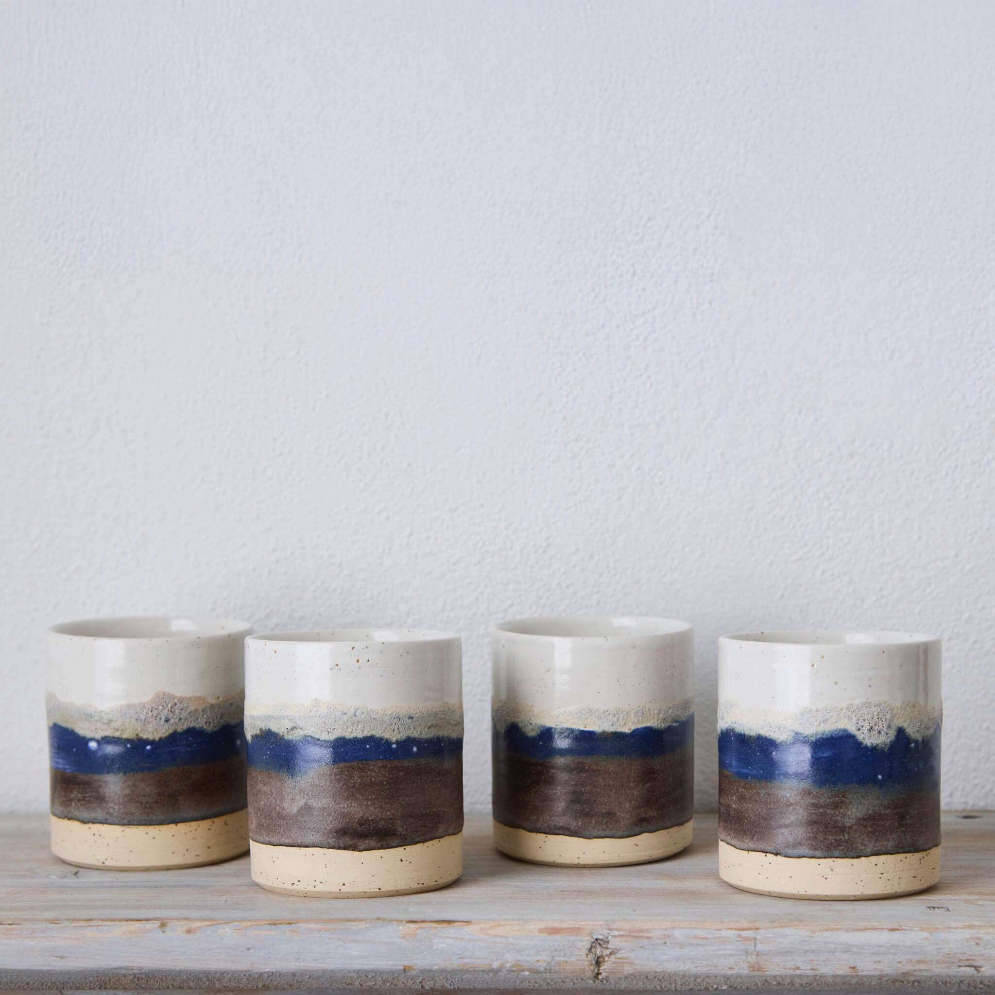Four ceramic cups with blue and brown stripes on a wooden surface against a light gray background