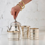 Three silver cups and a silver sugar bowl with a hand holding the sugar bowl lid against a white textured background