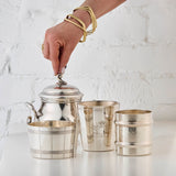 Three silver cups and a silver sugar bowl with a hand holding the sugar bowl lid against a white textured background