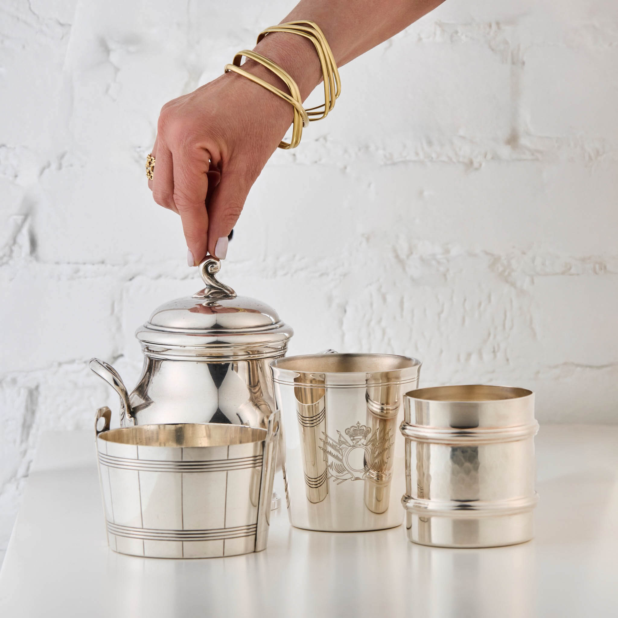Three silver cups and a silver sugar bowl with a hand holding the sugar bowl lid against a white textured background