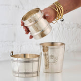 Hand holding a silver cup next to another silver cup and a silver ice bucket on a white background