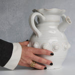 White ceramic pitcher with decorative elements held by a hand against a gray background