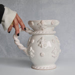 White ceramic vase with decorative patterns and a hand holding its handle against a gray background