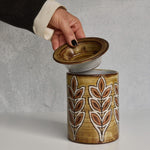 Ceramic Malarmey lidded jar with leaf pattern being opened by a hand on a gray background