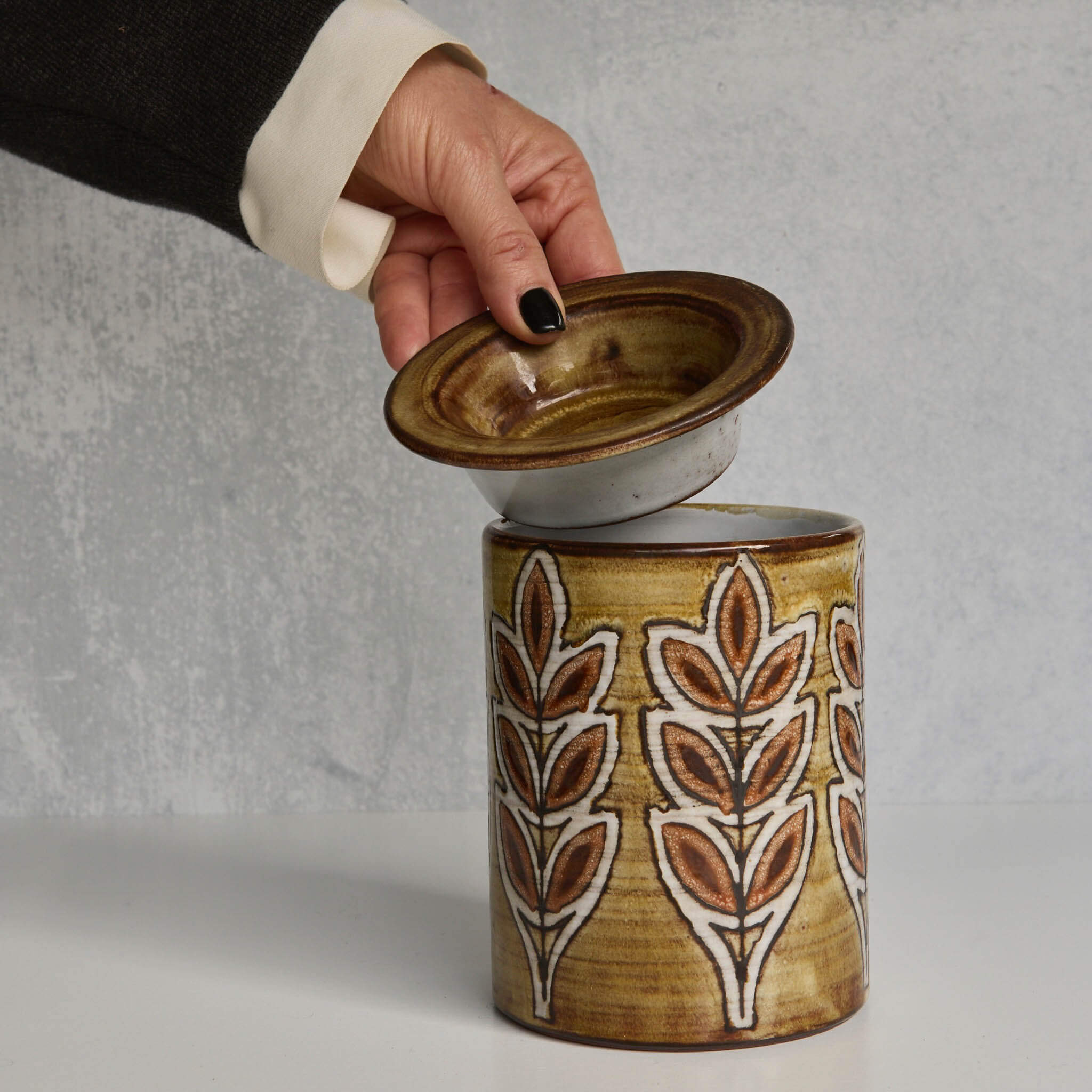 Ceramic Malarmey lidded jar with leaf pattern being opened by a hand on a gray background