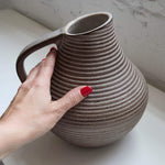 Hand holding a textured stoneware pitcher against a neutral background