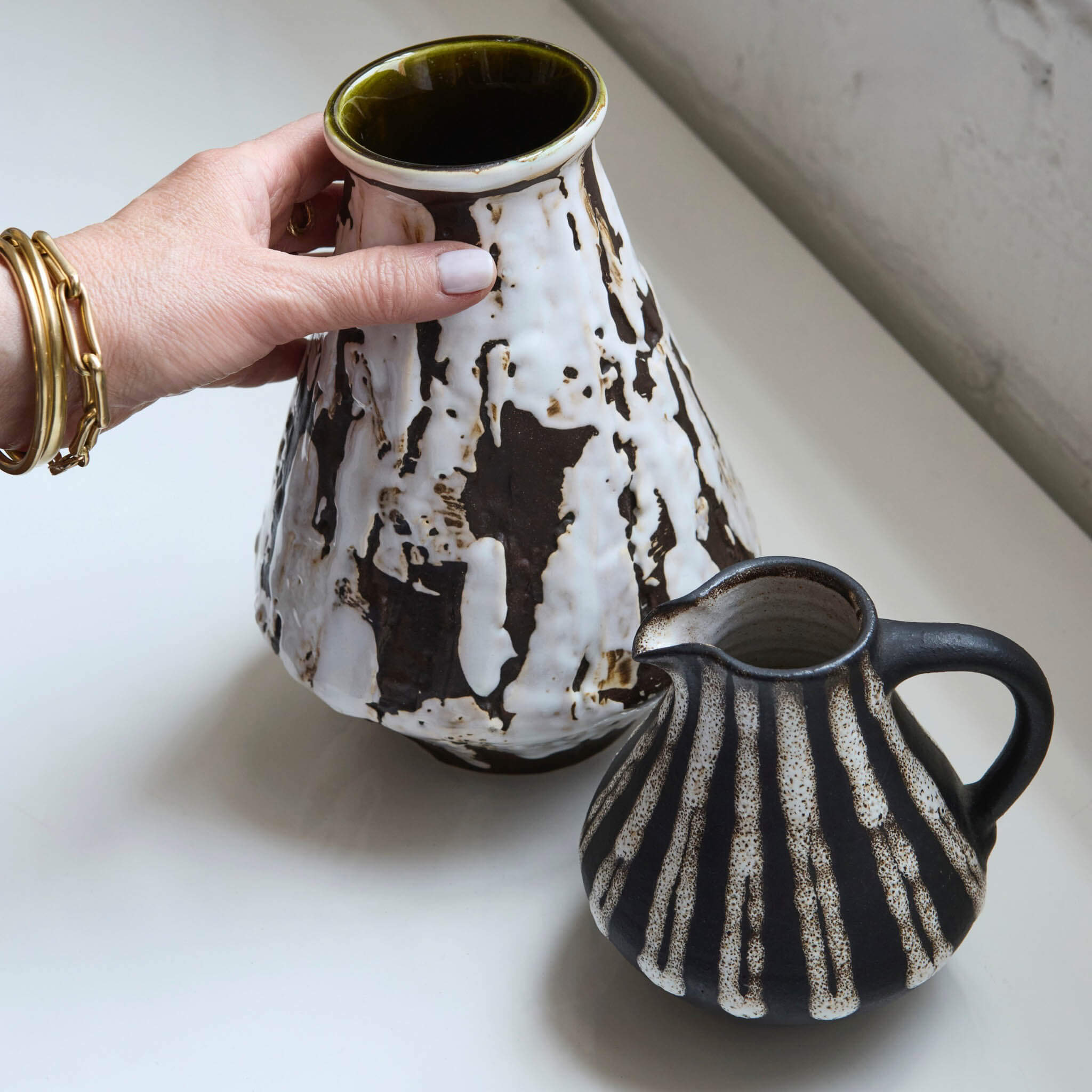 Hand holding a ceramic pitcher with another ceramic pitcher on a light surface.